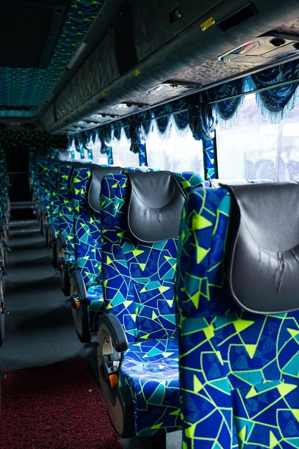 Interior of a bus with patterned seats featuring blue and green geometric designs, arranged in rows with visible windows.