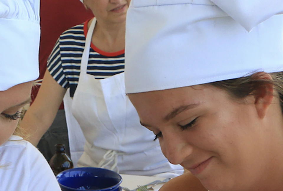 Two women in white chef hats and aprons chop herbs on a cutting board during a cooking class, with others in the background.