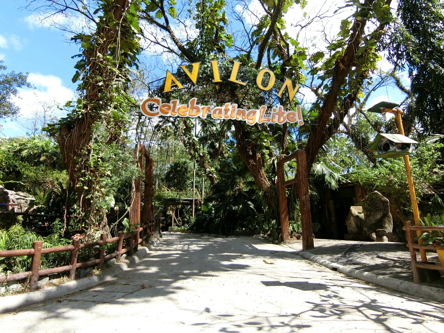 Entrance to a nature-themed area marked with a sign reading 'Avilon Celebrating Life!' surrounded by trees and foliage.