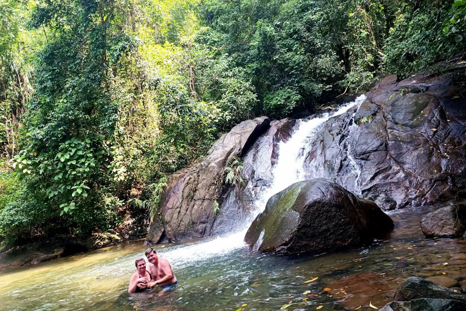 Two people standing in a natural pool near a waterfall surrounded by lush greenery and rocks.