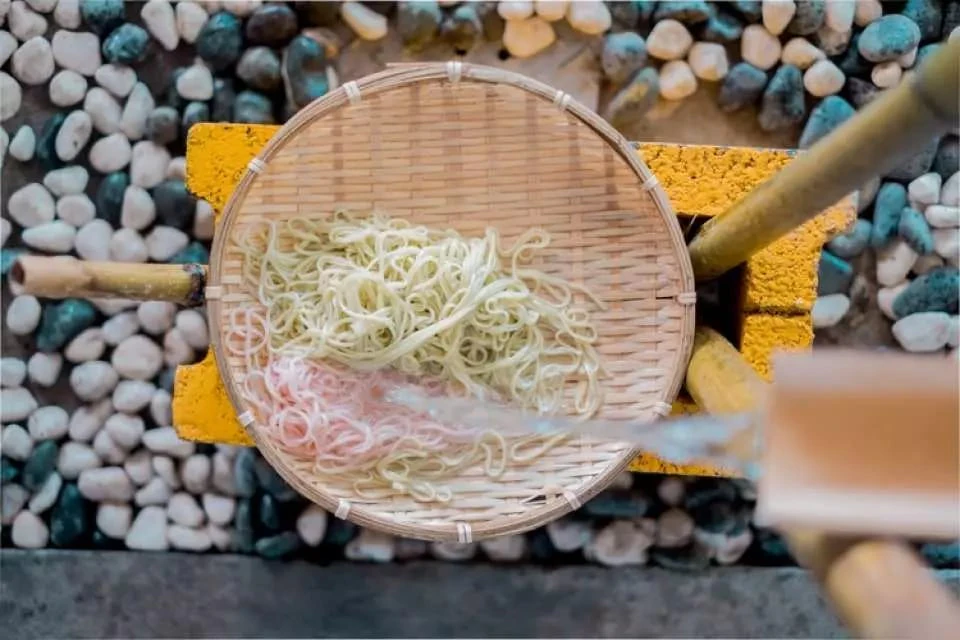 A bamboo basket containing yellow and pink noodles placed on a yellow surface, surrounded by pebbles.