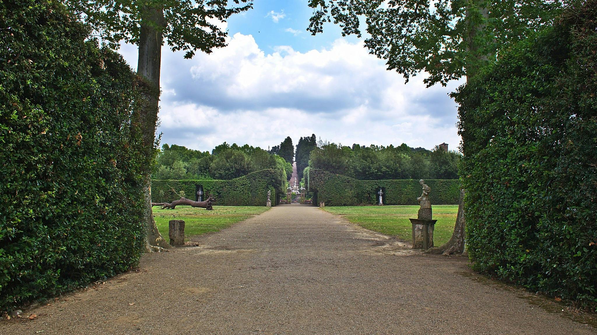 A wide gravel path flanked by statues, leading through a manicured garden with tall hedges, trees, and a cloudy sky above.