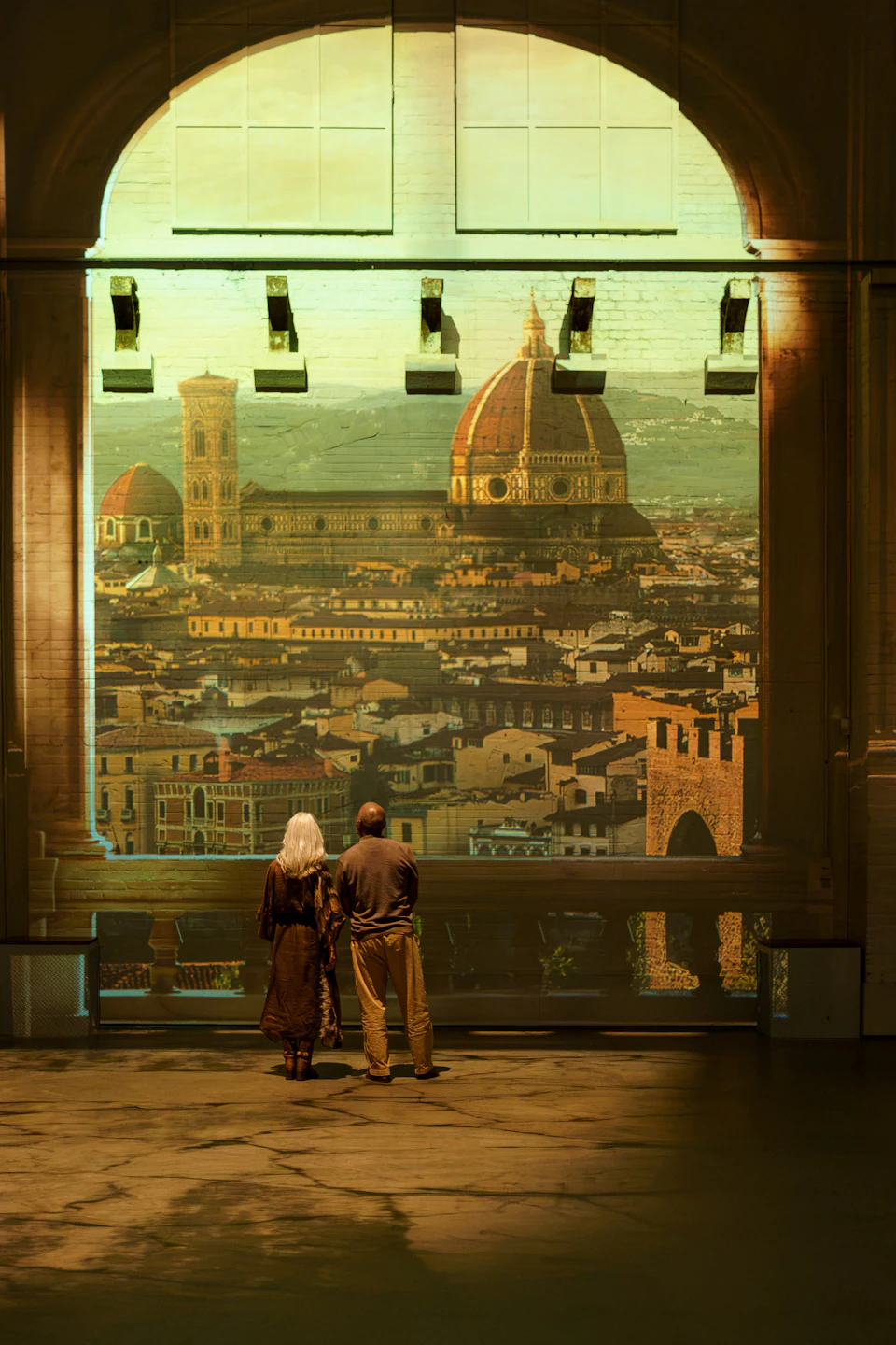 Two people stand facing a large illuminated image of Florence, featuring the Duomo and cityscape.