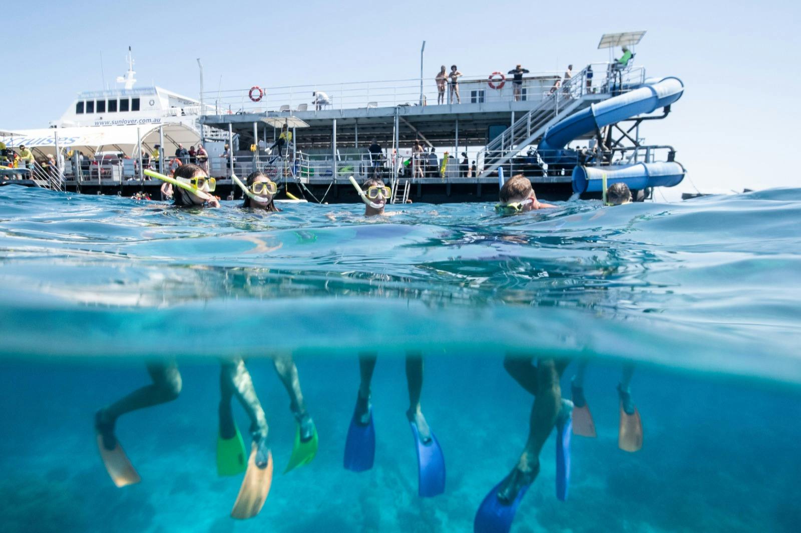 Persone che fanno snorkeling in un'acqua limpida e blu vicino a un molo con scale e uno scivolo, indossando pinne e maschere da snorkeling.