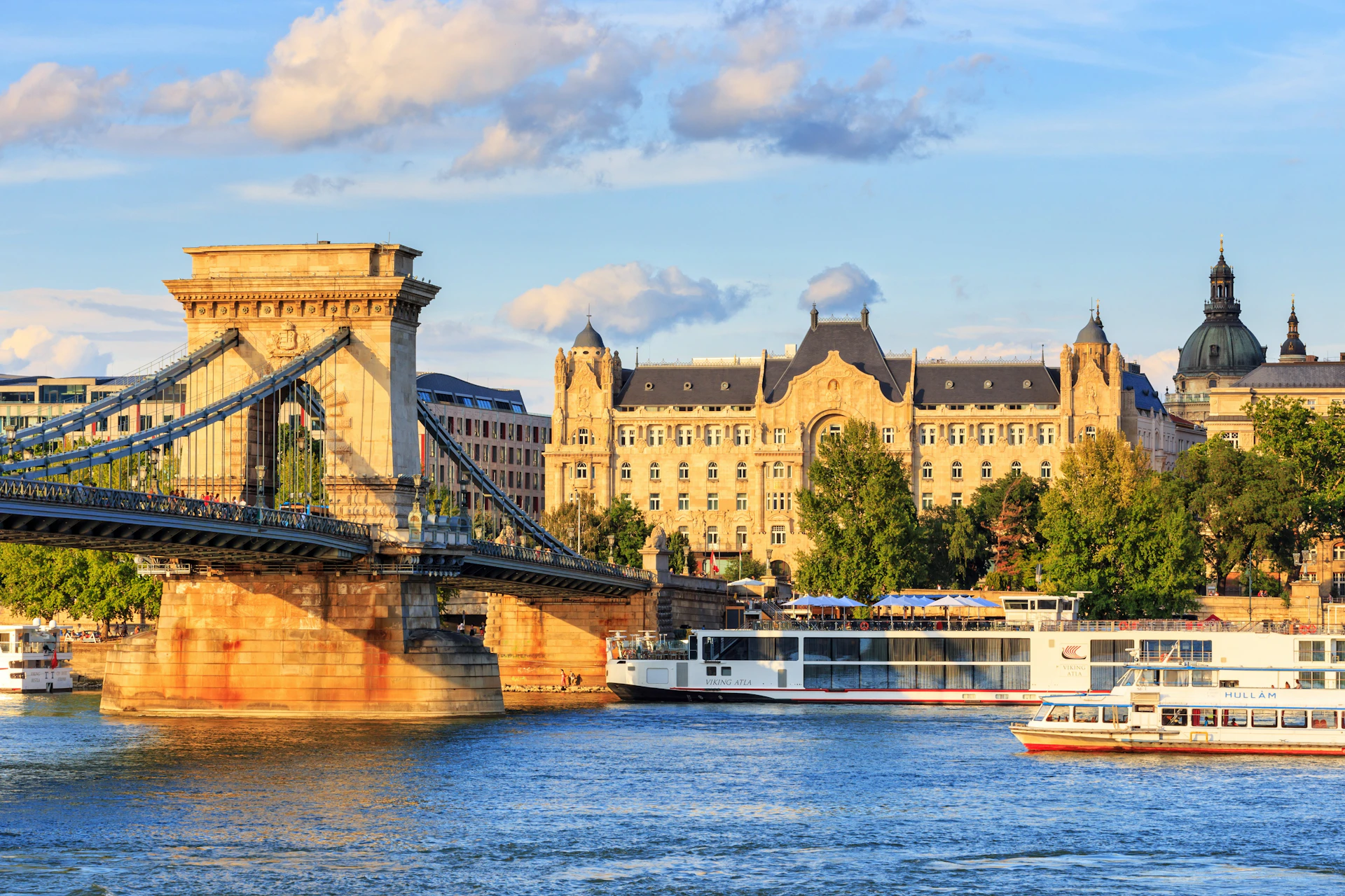 A scenic view of the Chain Bridge and a historic building in Budapest, with boats on the Danube River under a sunny sky.
