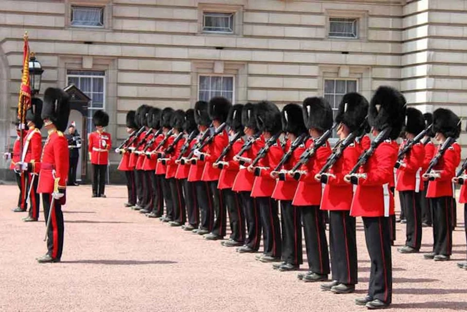 A row of guards in red uniforms and black bearskin hats stand in formation outside a stone building with multiple windows.