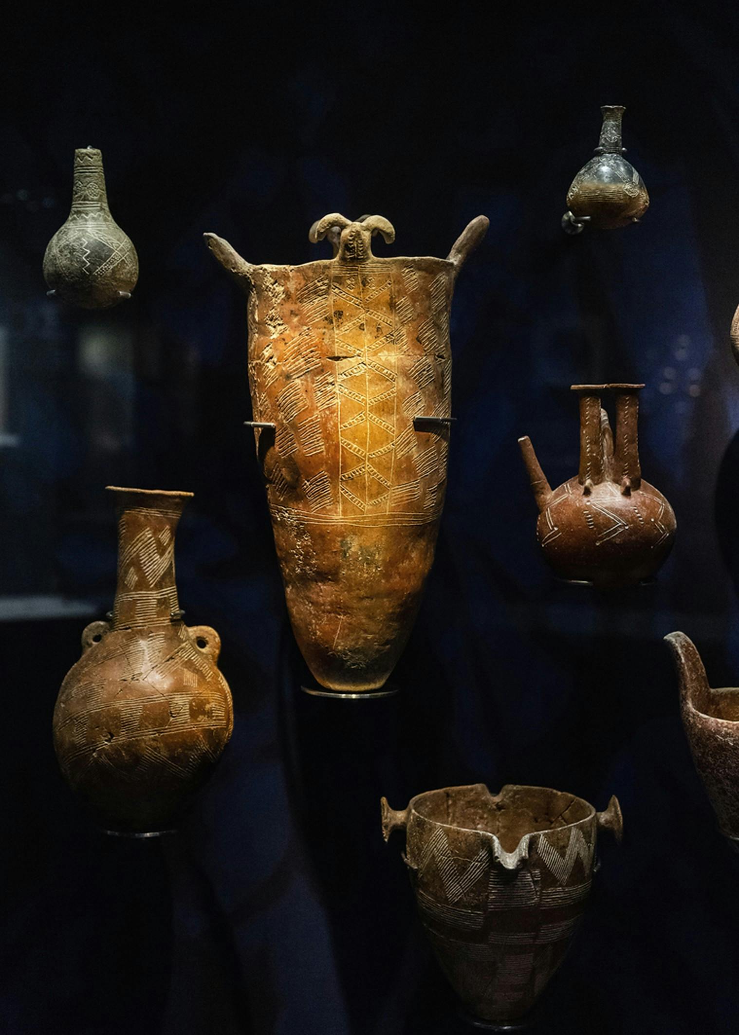 A display of various ancient pottery pieces with intricate patterns, including bowls, jars, and a tall vessel, against a dark background.