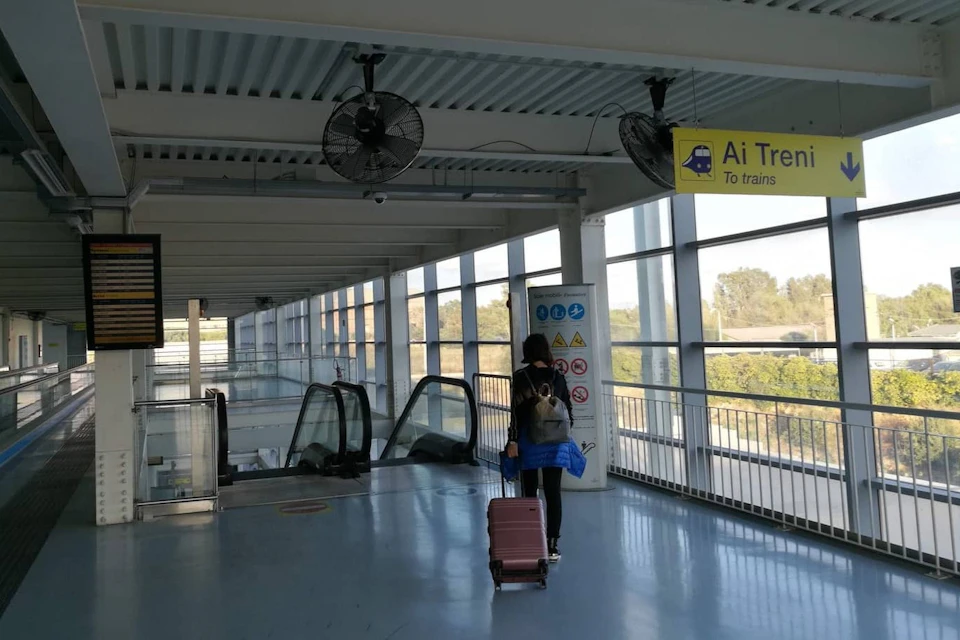 Person with a suitcase walking toward escalators in a spacious indoor area. Sign overhead reads "Ai Treni - To trains."
