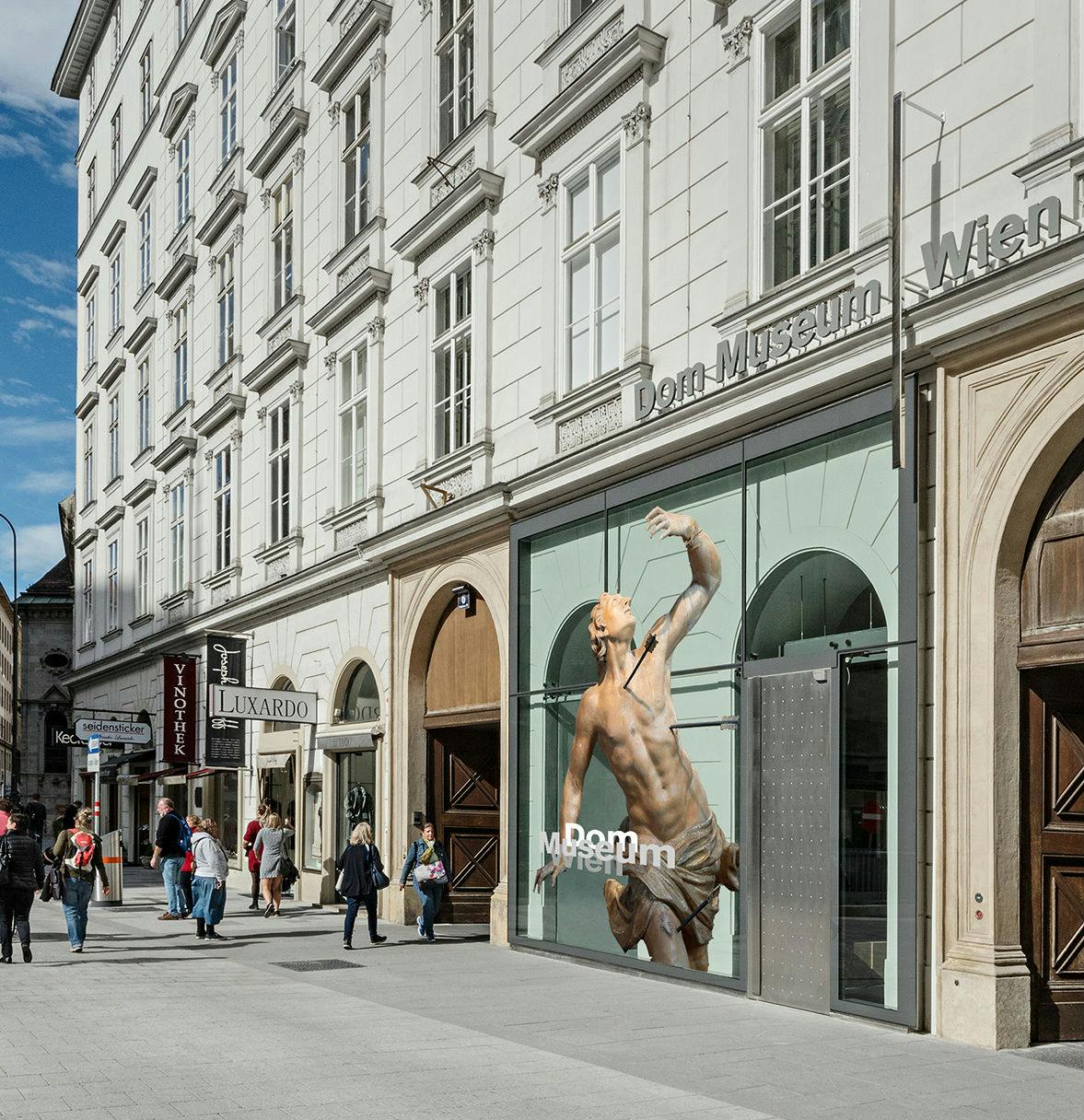 Gente paseando junto al Museo Dom de Viena, que exhibe una gran escultura de un hombre en el escaparate. La calle está repleta de tiendas.