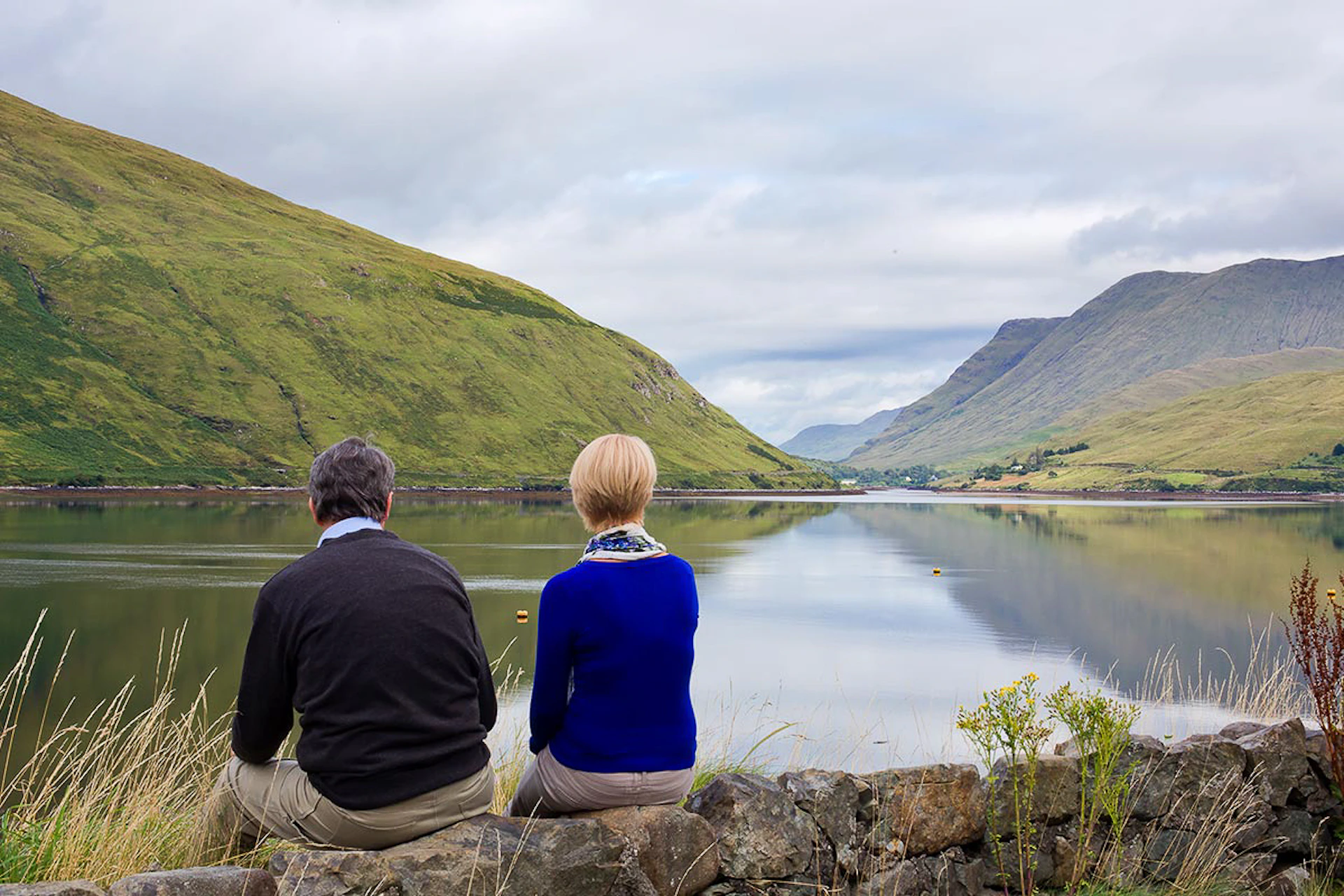 A man and woman sit on rocks by a calm lake surrounded by green hills and mountains under a cloudy sky.