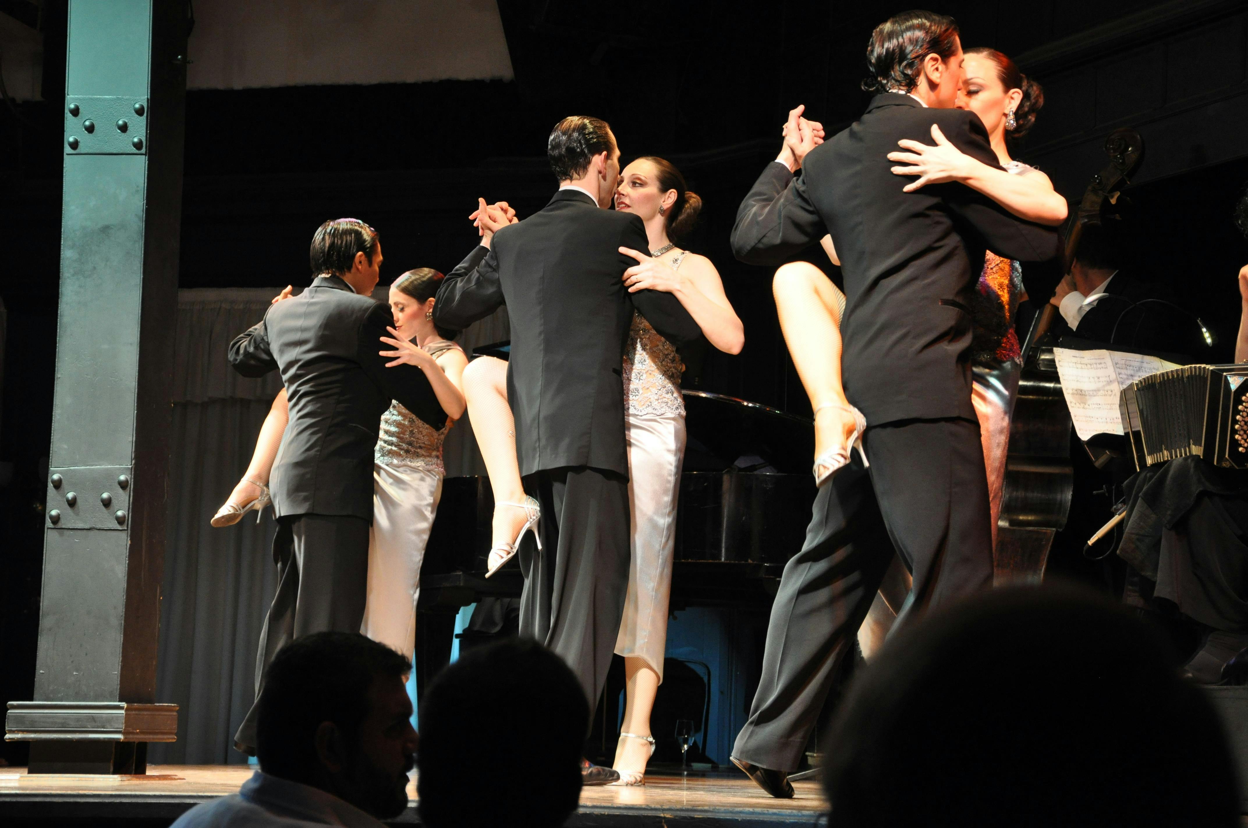 Three couples perform elegant dance moves, wearing formal attire, in a dimly lit venue with musicians in the background.