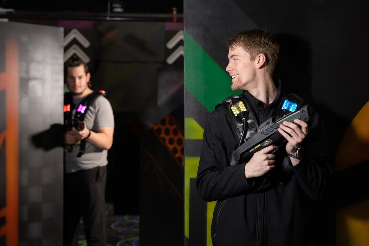 Person holding a laser tag gun and wearing a laser tag vest, standing near dark, neon-lit walls.