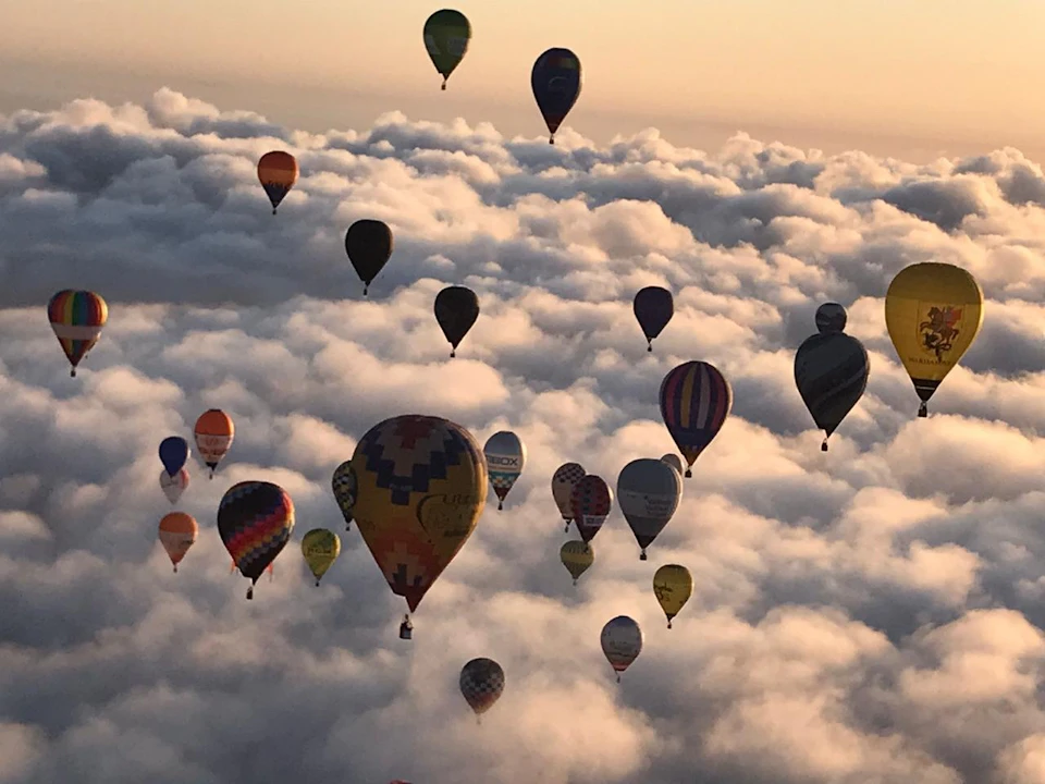 Numerous colorful hot air balloons float above a sea of clouds during a sunrise or sunset.