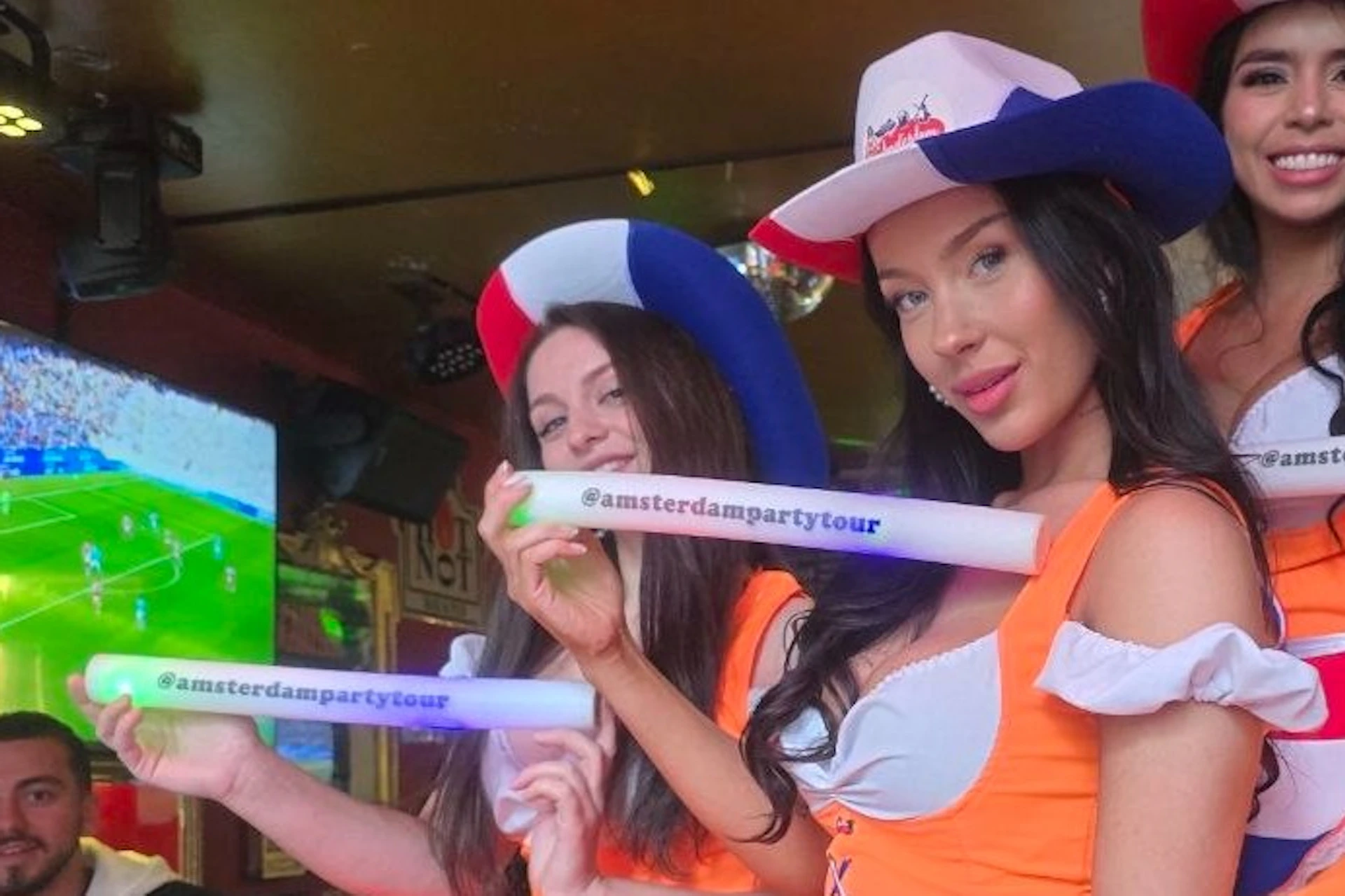 Three women in orange dresses and themed hats hold promotional items in a lively indoor setting with a soccer match on TV.
