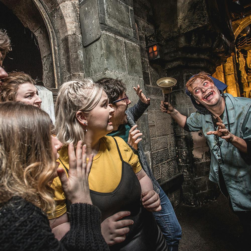 A group of people react with fear in front of a spooky, costumed character holding a bell in a dimly lit, medieval-looking alleyway.