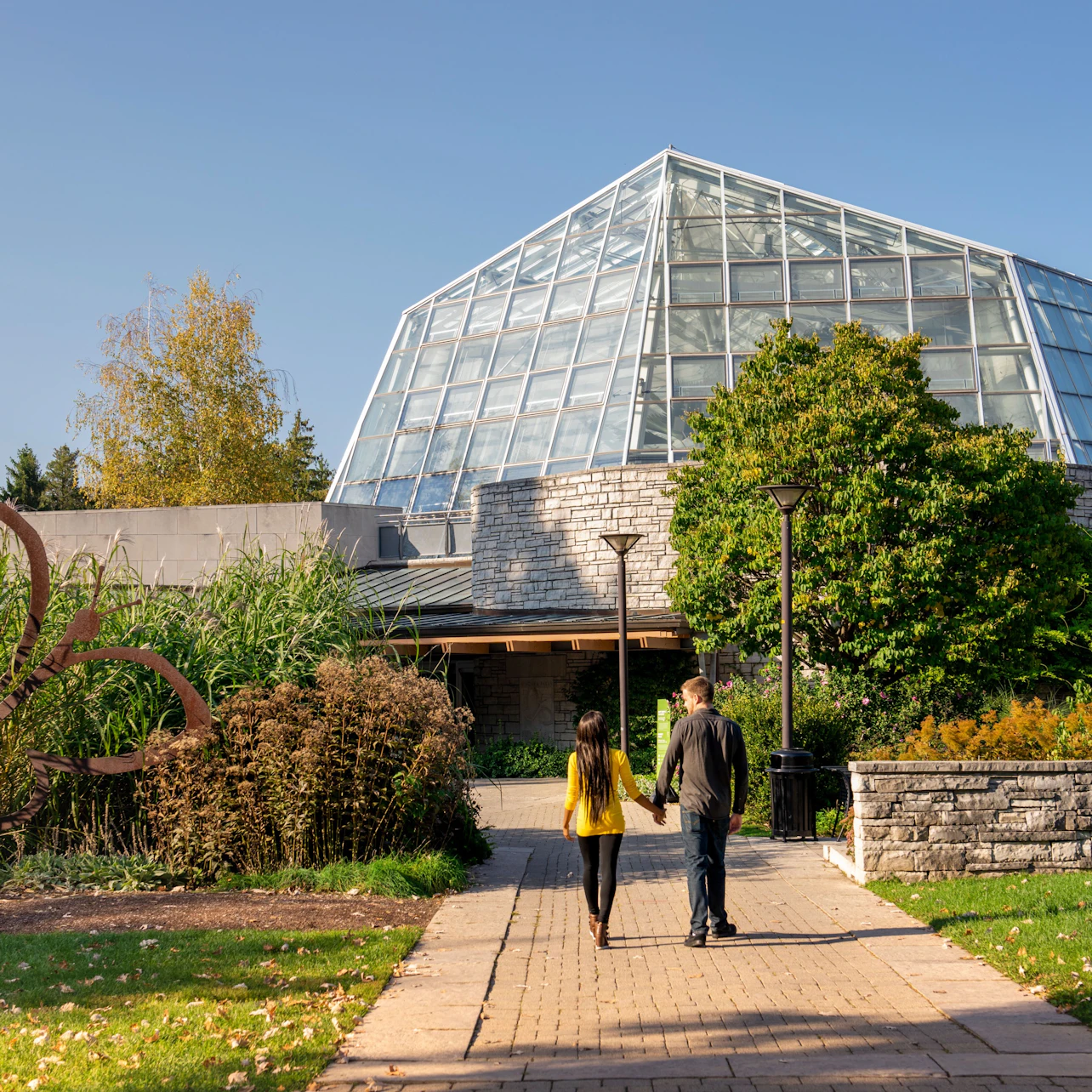 Two people walking on a paved path towards a modern glass building surrounded by trees and greenery on a sunny day.