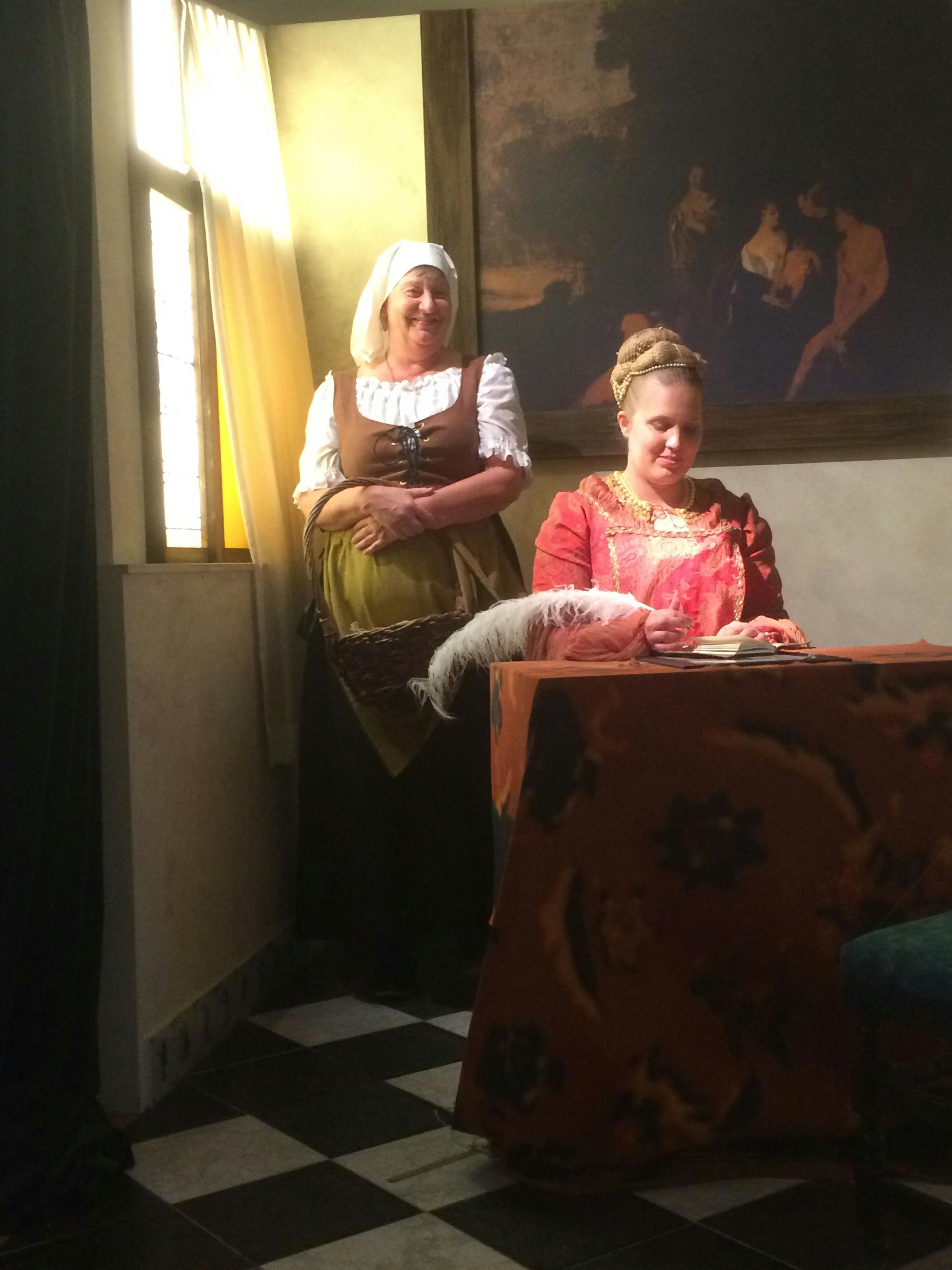 Duas mulheres em trajes da época, numa sala pouco iluminada, uma sentada a uma mesa, a escrever com uma pena, e a outra de pé, a sorrir.