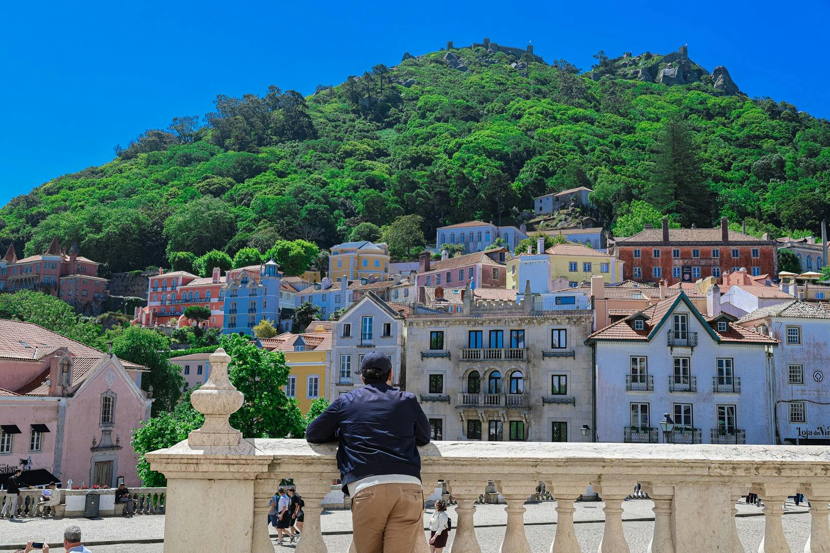 A person leans on a stone balustrade, overlooking colorful buildings and a wooded hill under a clear blue sky.
