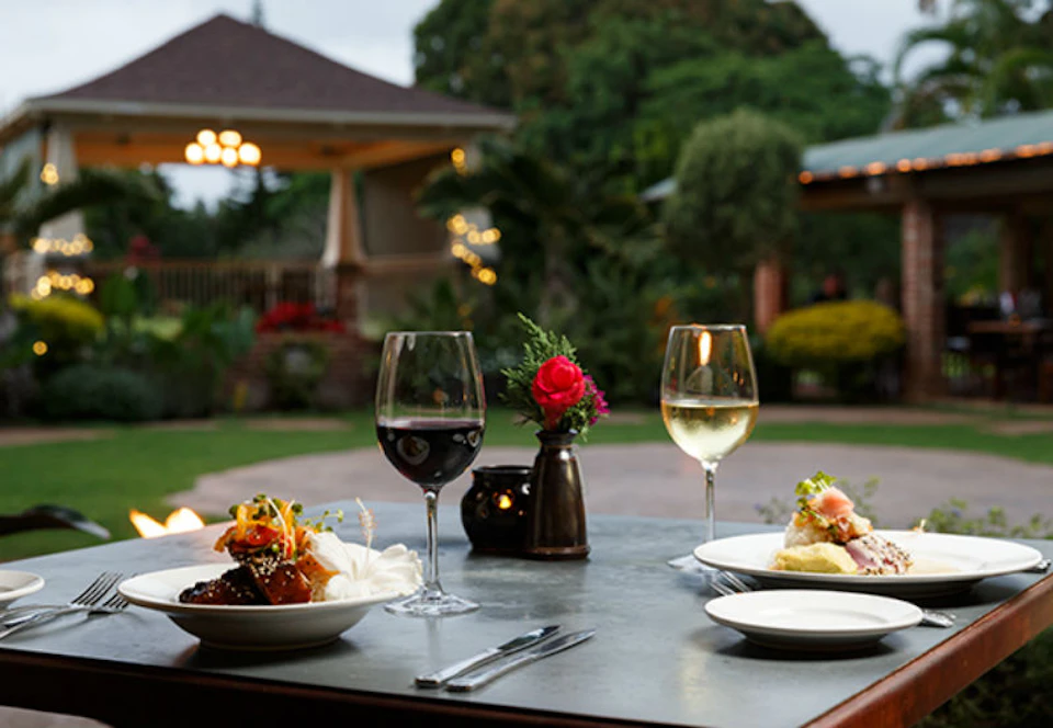 Outdoor dining table with two wine glasses, plates of food, and a small vase with a red flower. Blurred garden background.