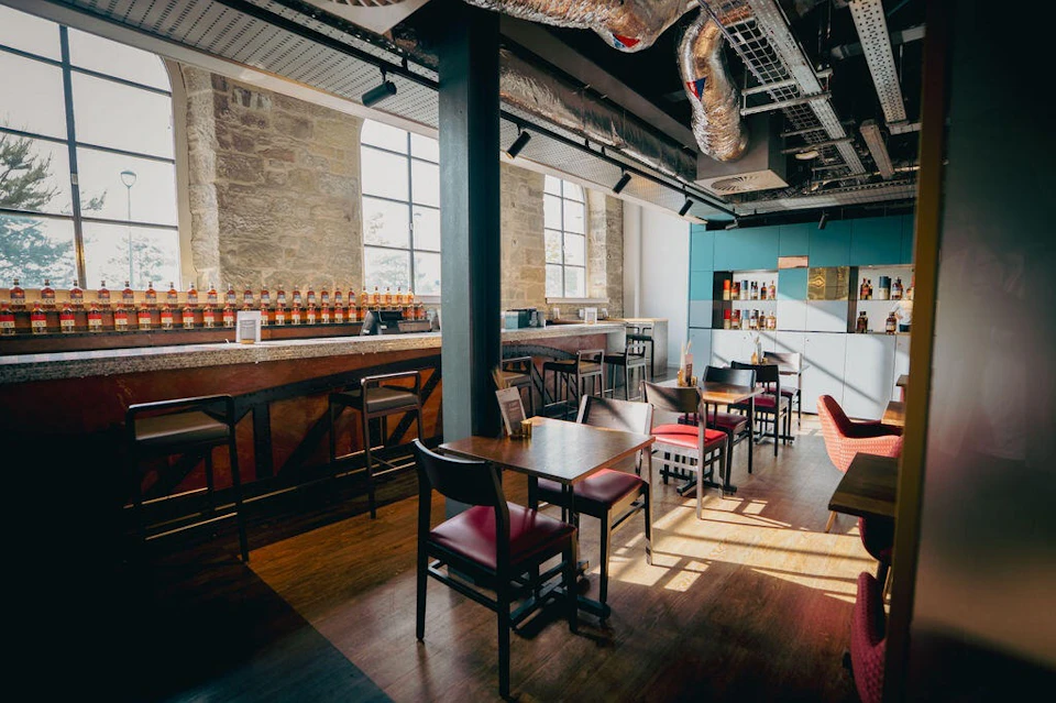 A modern cafe with wooden tables, chairs, a bar with bottles, large windows, and industrial-style decor.