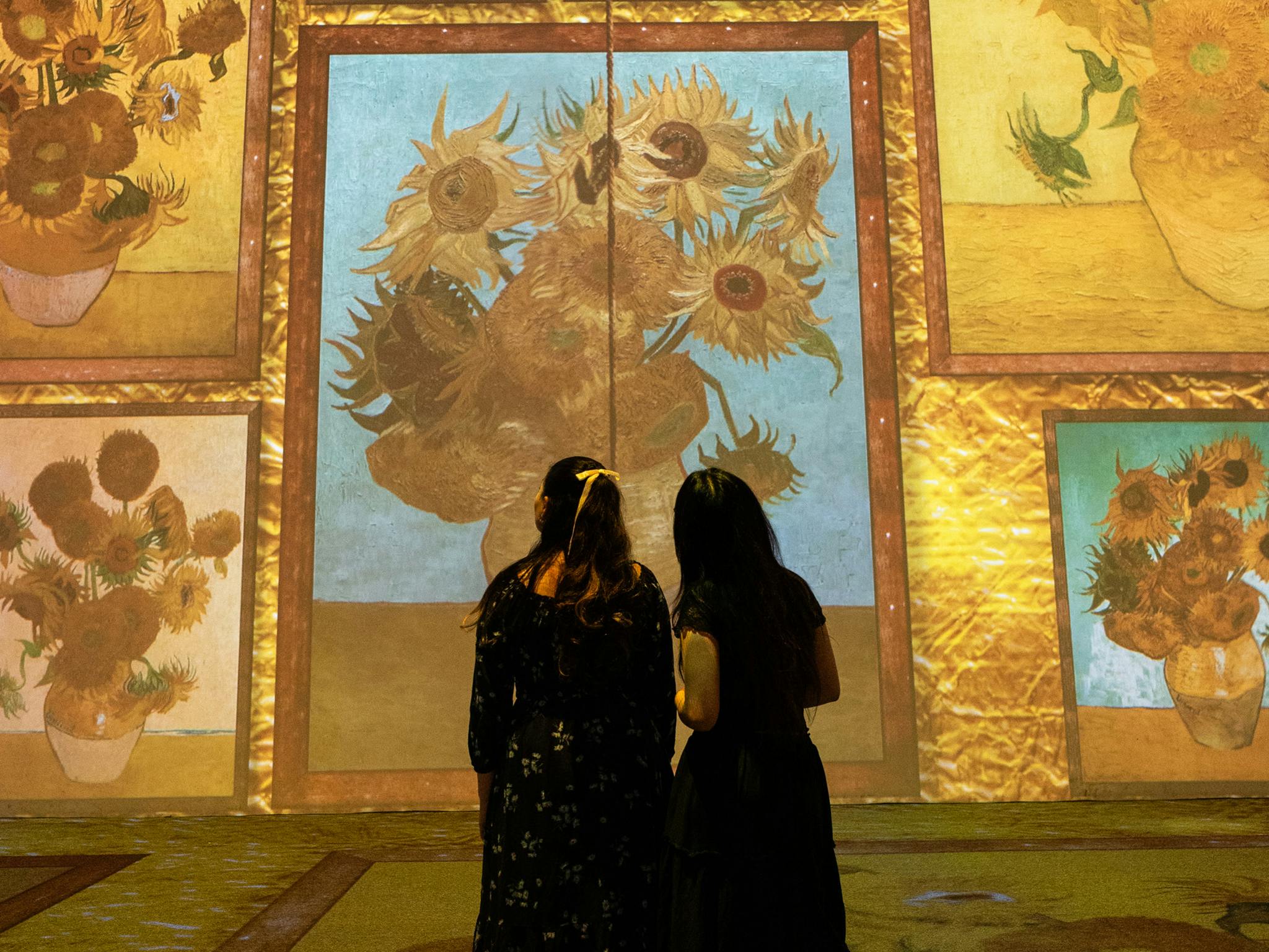两位女士站在梵高向日葵画作的大型数码展览前，欣赏这件艺术品。