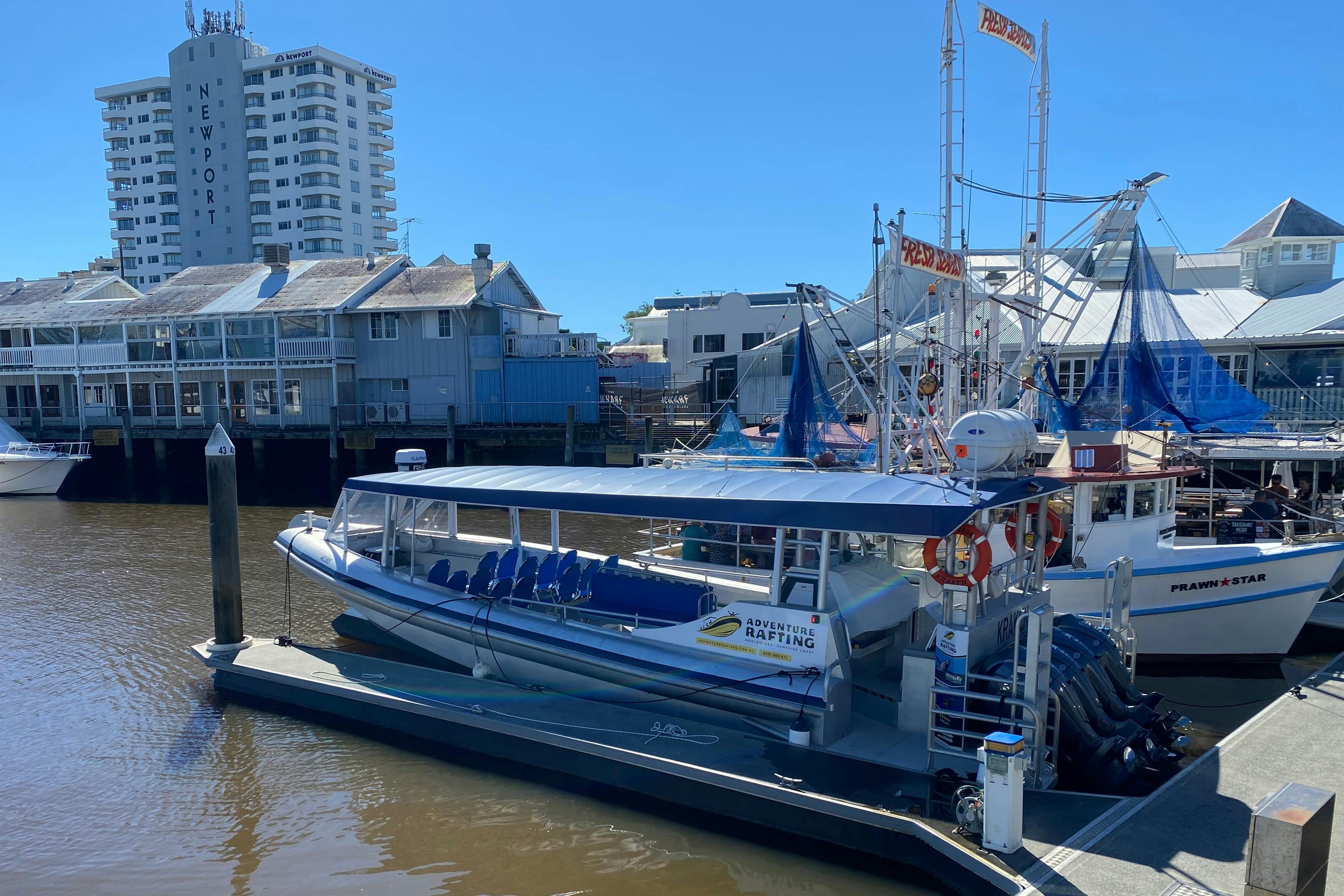 Een klein rondvaartbootje ligt aangemeerd aan een steiger, omringd door andere boten, met gebouwen en een heldere lucht op de achtergrond.