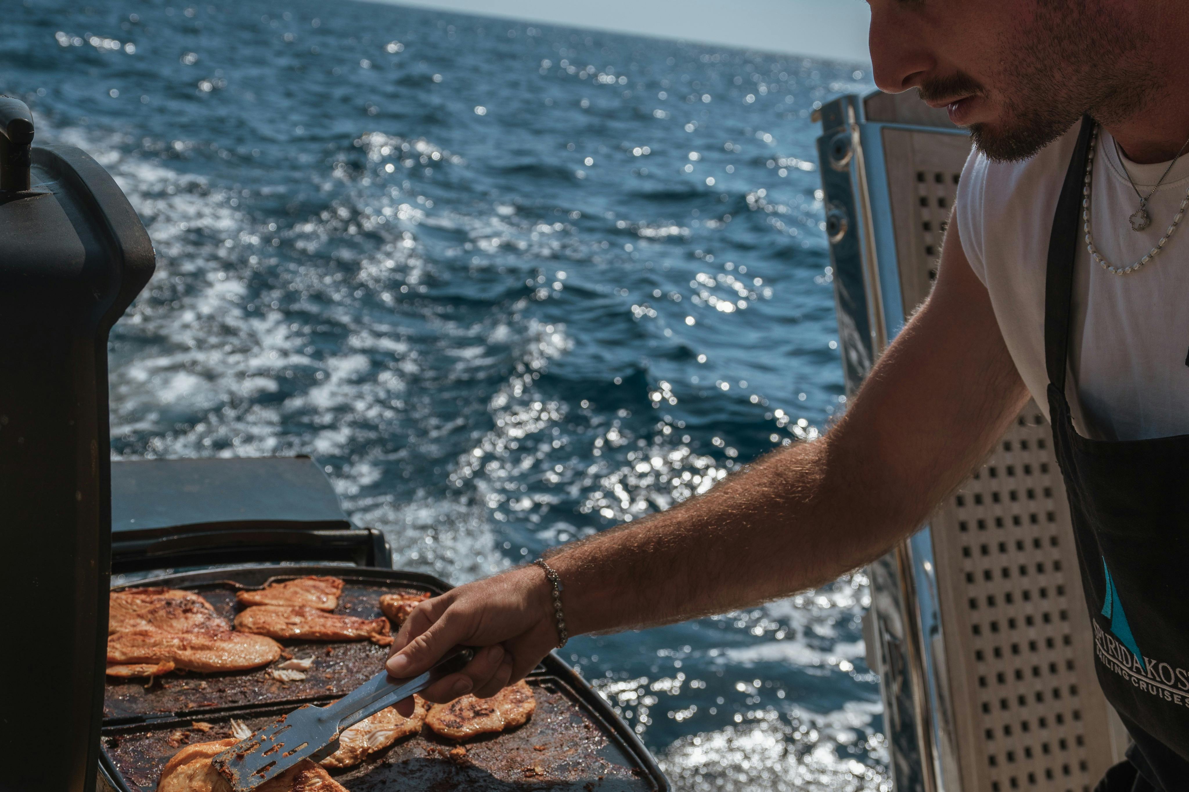 A person grilling food on a barbecue with the ocean visible in the background on a sunny day.