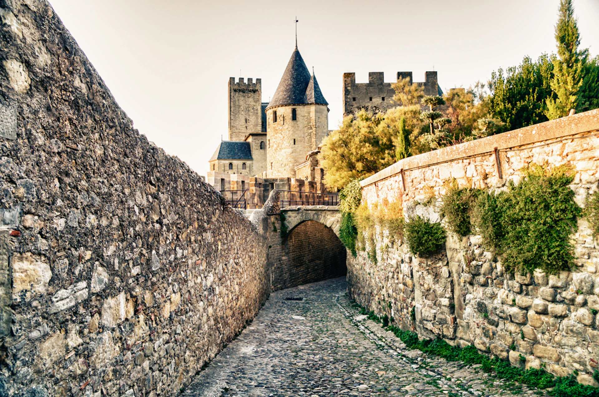 A cobblestone path leads to a historic stone castle with multiple towers and a bridge, flanked by high stone walls.