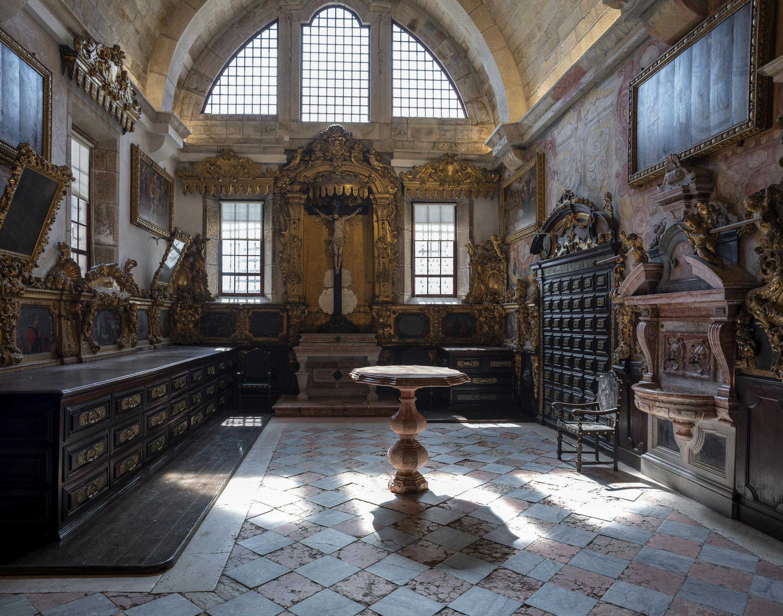 Ornate room with arched windows, intricate wooden furniture, a central pedestal table, and sunlight streaming onto a checkered floor.