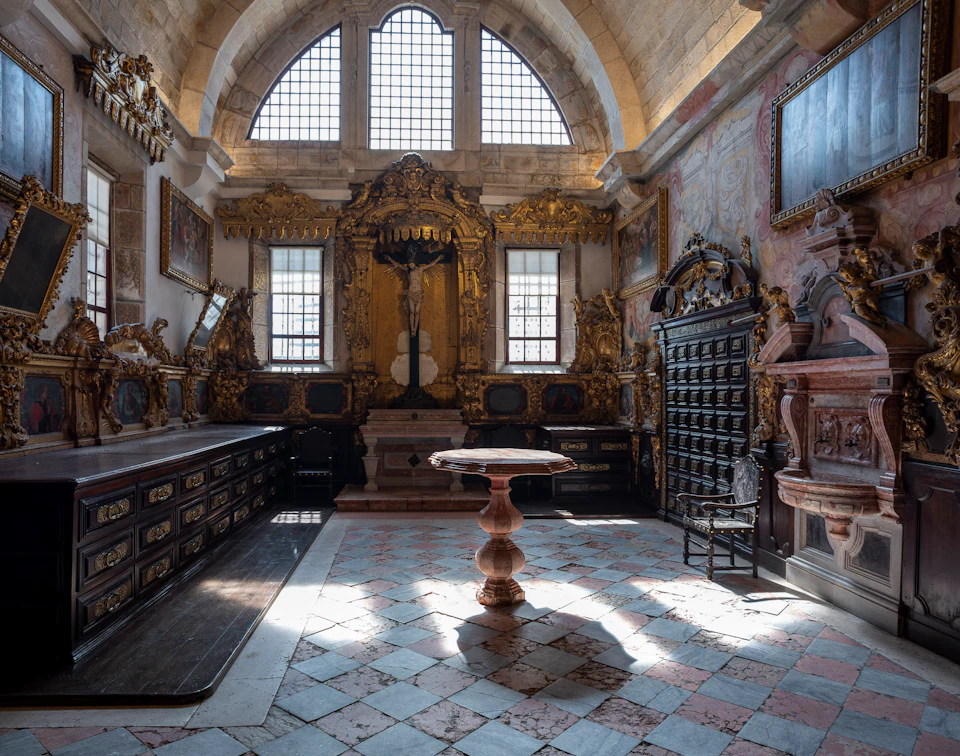 An ornate, sunlit room with high arched windows, golden altar, decorative wall carvings, and a central wooden pedestal table.
