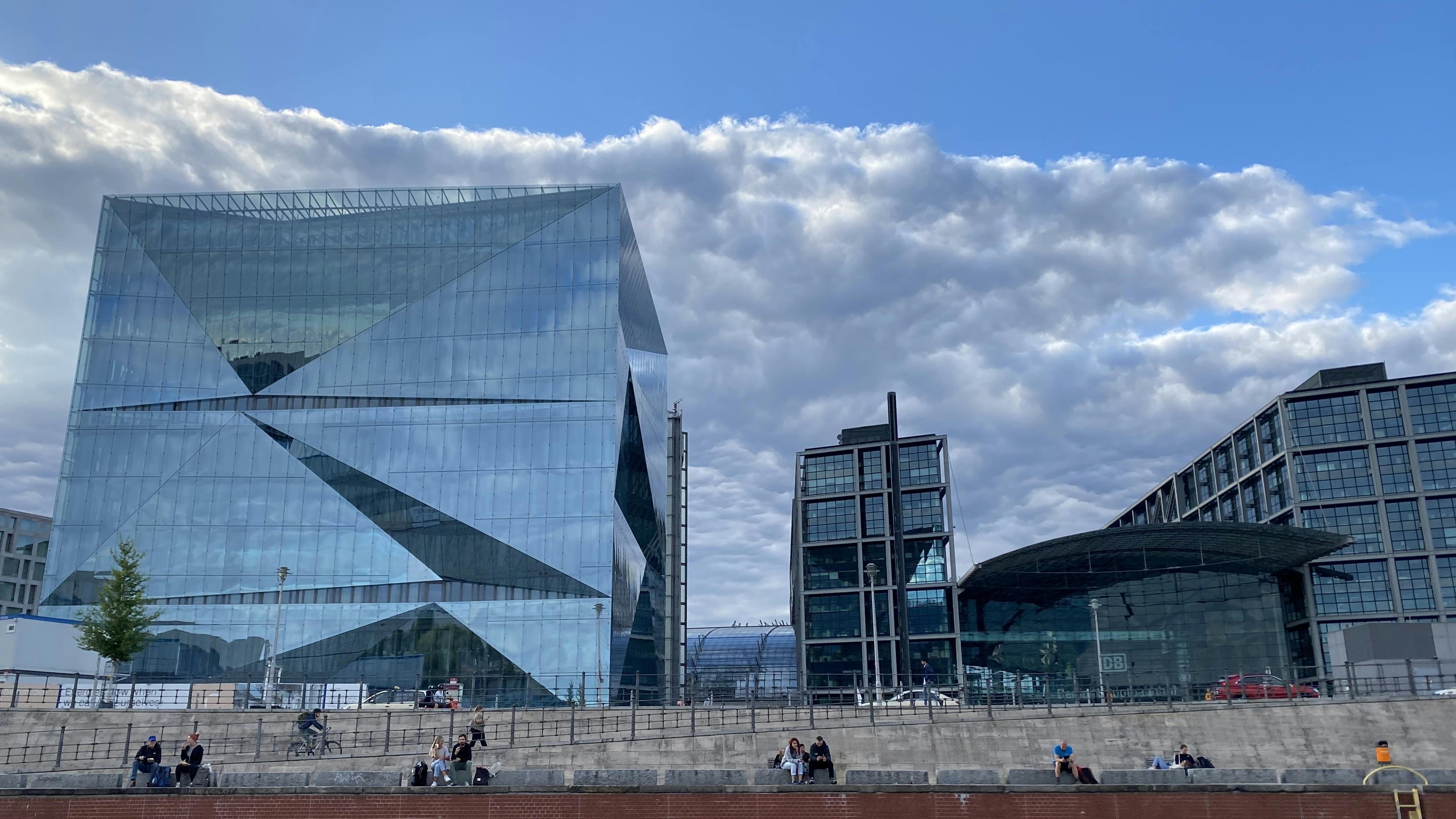 Σύγχρονα γυάλινα κτίρια με φόντο ένα συννεφιασμένο γαλάζιο ουρανό, με τους ανθρώπους να κάθονται σε σκαλοπάτια στο προσκήνιο.