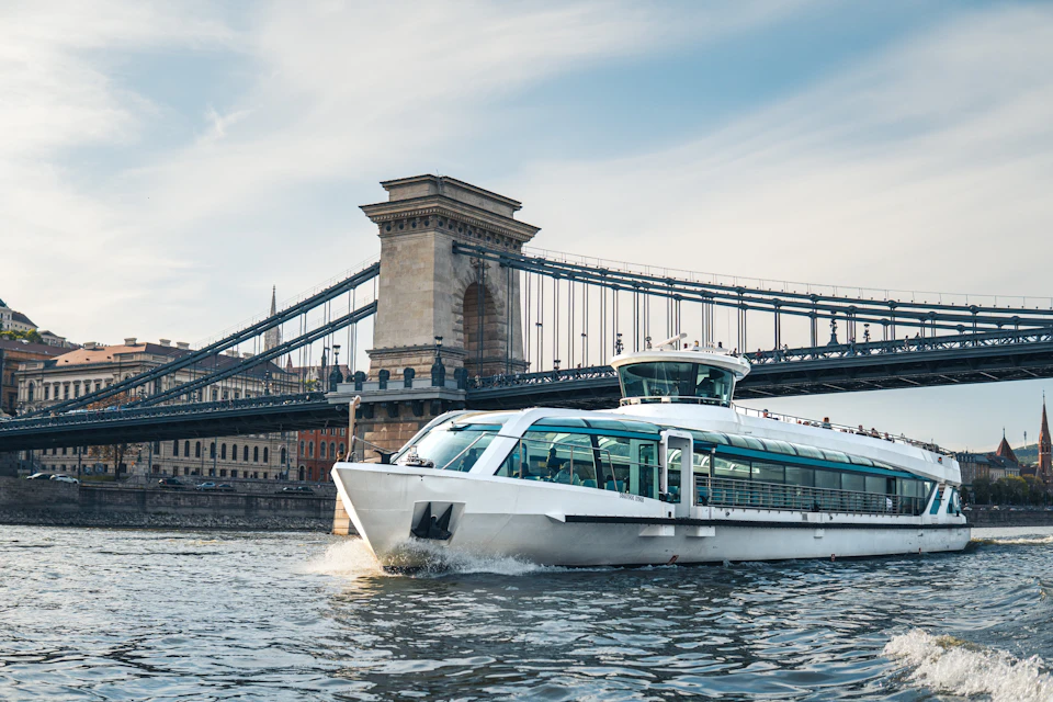A white boat cruising on a river, with a large suspension bridge and urban buildings in the background under a cloudy sky.