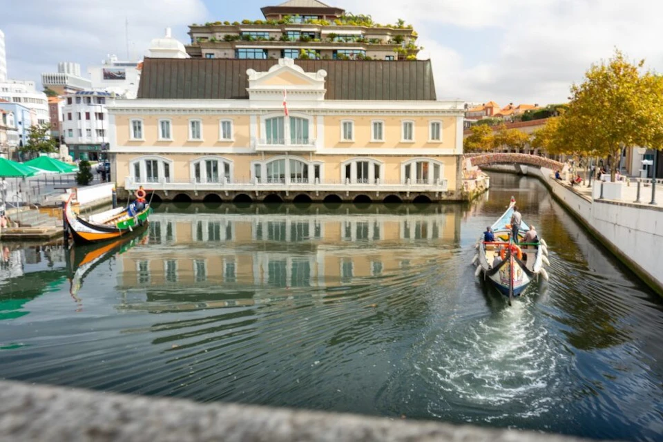 Two boats move along a calm canal in front of a ornate, beige building with arched windows. Trees and other buildings are visible in the background.