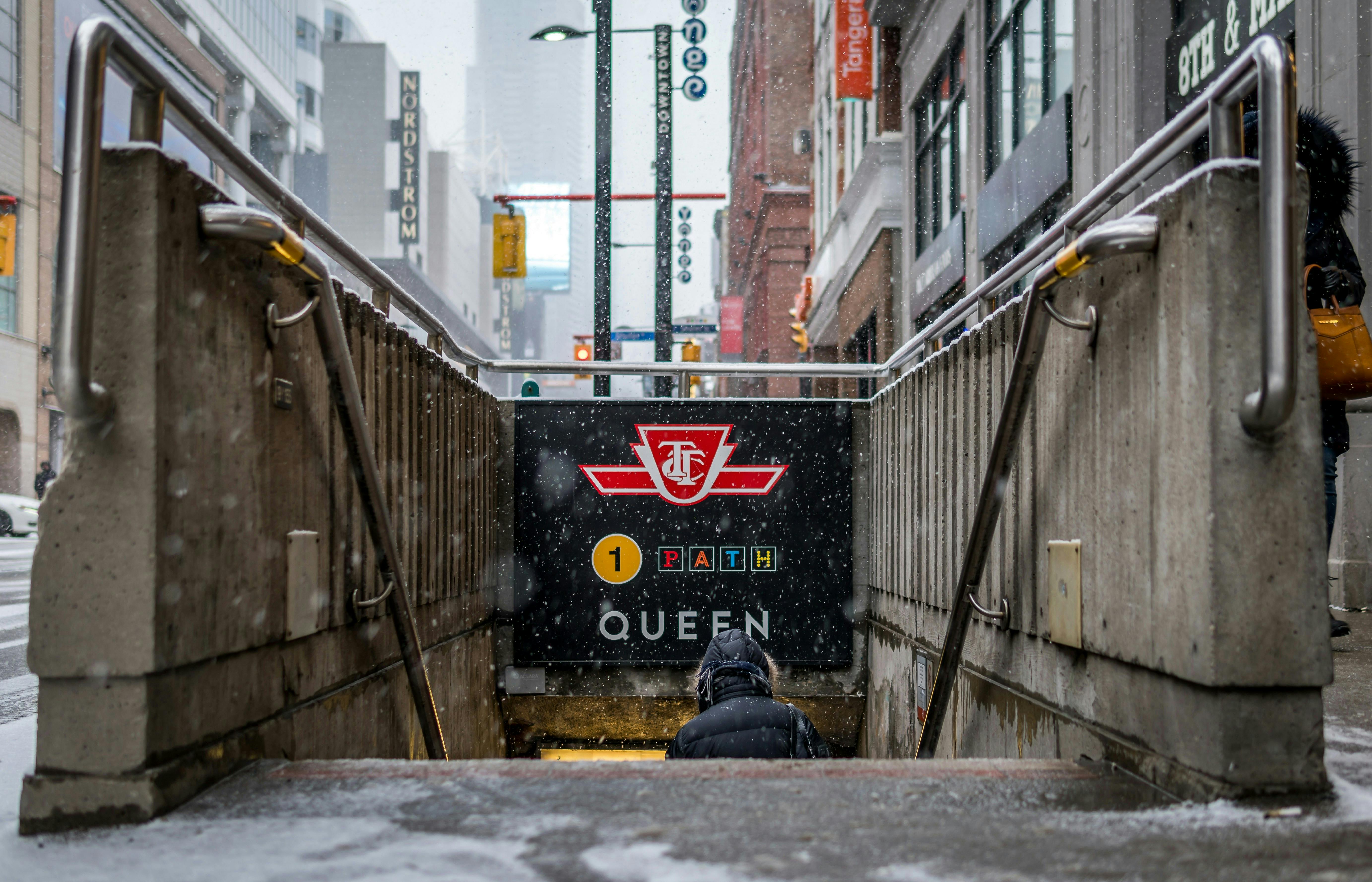 Entrée de métro avec le logo de la TTC, le panneau "Queen", la neige qui tombe et une personne qui descend les escaliers dans un environnement urbain.