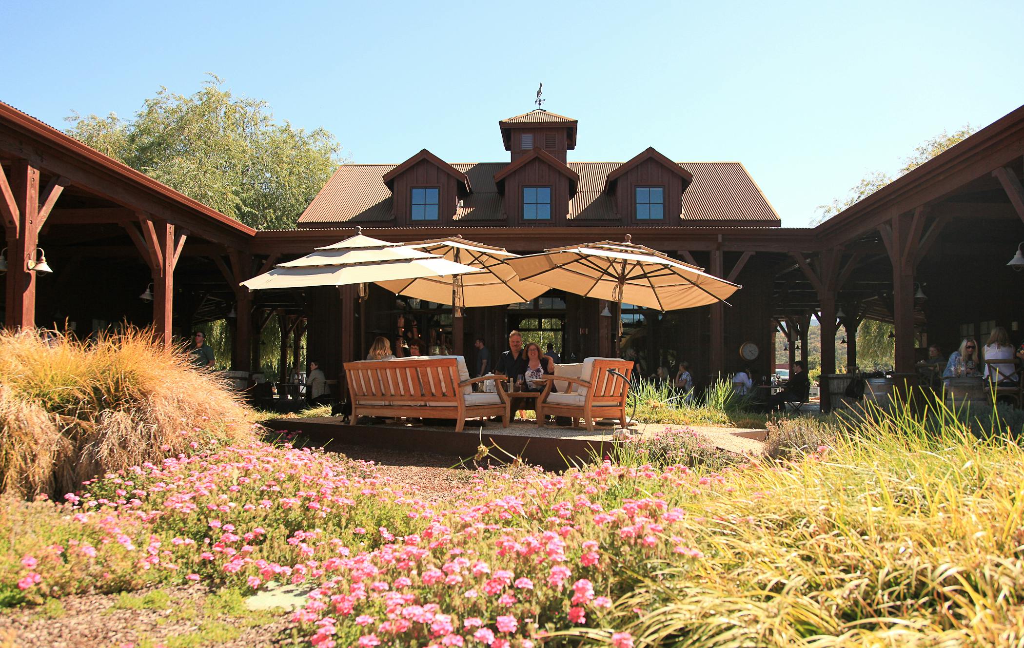 複数のドーマーの窓とピンクの花の庭がある素朴な建物の前のパティオの大きな傘の下に座っている人々。