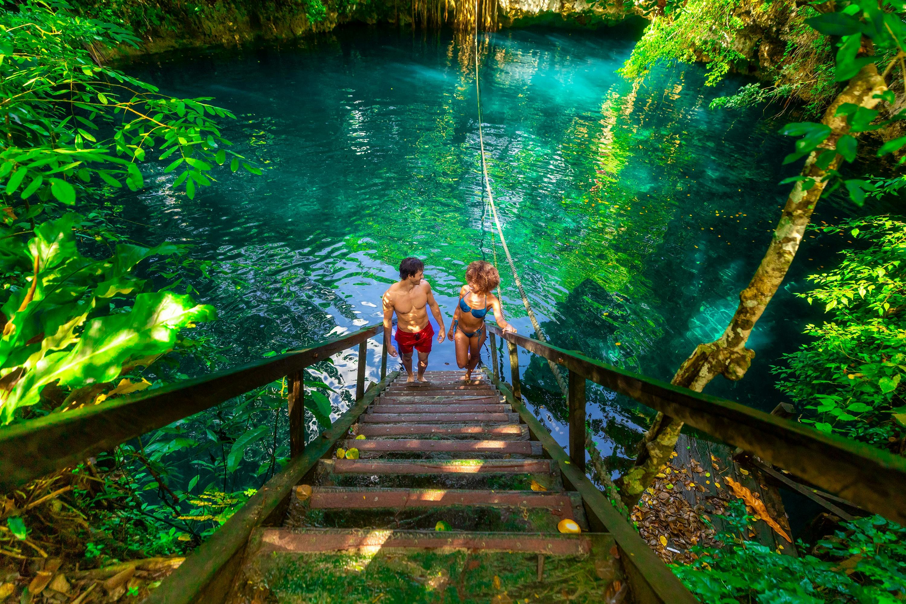 Two people in swimwear ascend wooden stairs from a clear turquoise natural pool surrounded by lush green foliage.