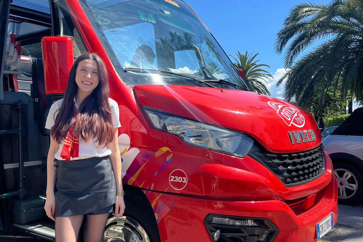 Smiling woman with long hair stands next to a red bus with colorful decals and the number 2303, under a clear blue sky.