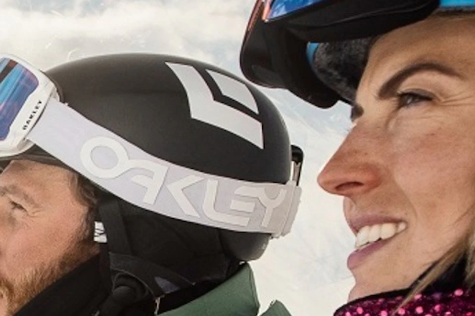 Three skiers in colorful outfits ride a ski lift on a sunny day, with snowy mountains and other skiers visible in the background.