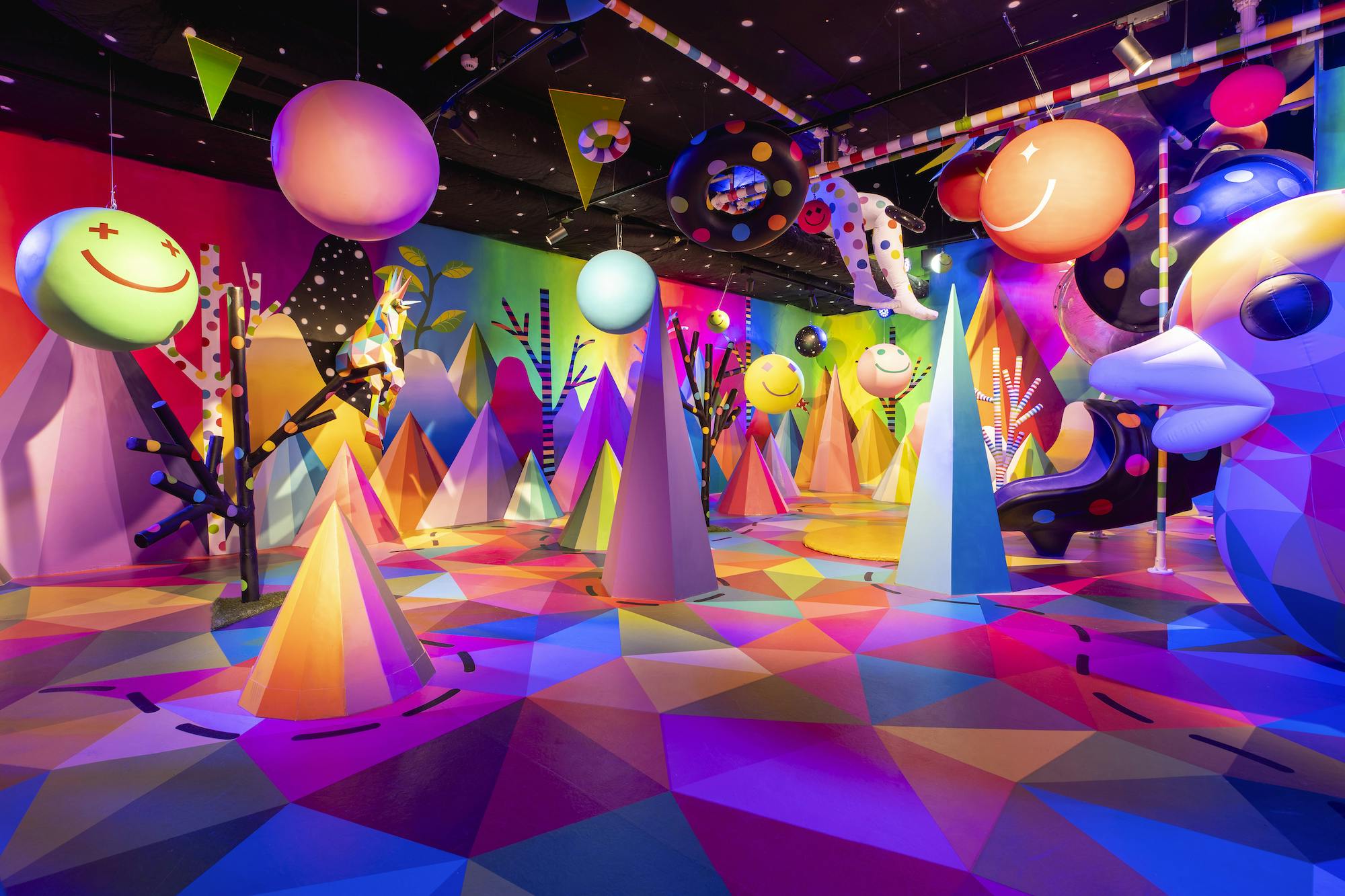 鮮やかな幾何学模様、円錐形の構造、吊り下げ式のスマイリーフェイス、水玉模様の要素、ライトアップされた木々を備えたカラフルな部屋。