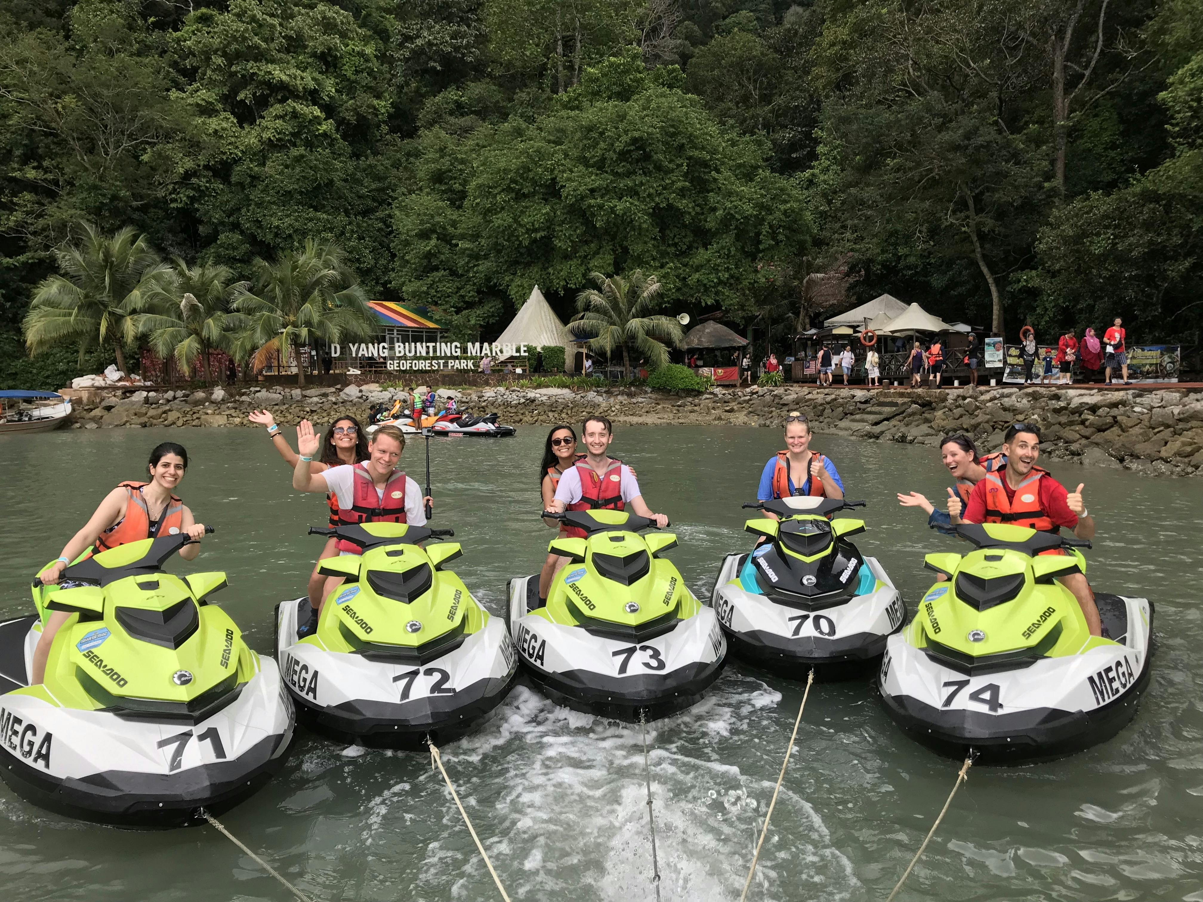 Cinq personnes sur des jet-skis dans un cadre tropical avec des arbres denses et un panneau en arrière-plan indiquant "Dayang Bunting Marble Geoforest Park" (Parc géoforestier de marbre de Dayang Bunting).
