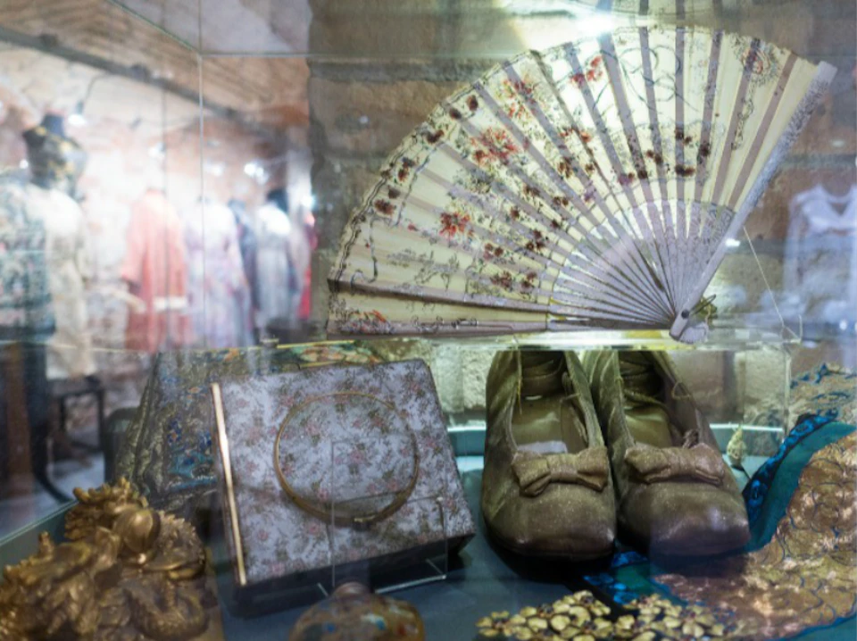 Display case containing a decorative fan, vintage shoes with bows, a floral handbag, and other antique accessories.