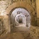 Ancient stone ruins with multiple arches, brick and stone masonry, dusty floor, and ambient light highlighting the structure.
