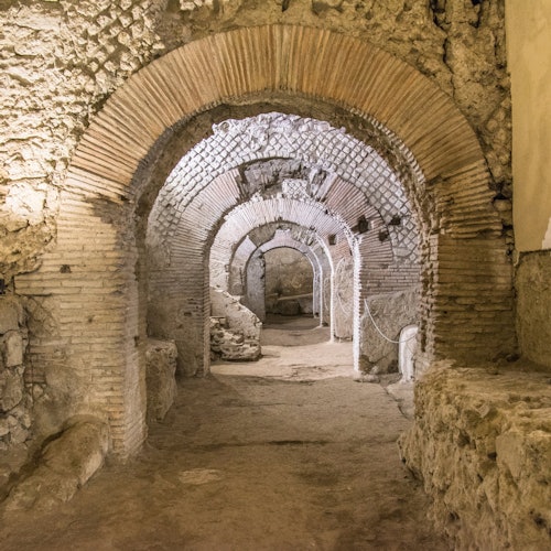Ancient stone ruins with multiple arches, brick and stone masonry, dusty floor, and ambient light highlighting the structure.