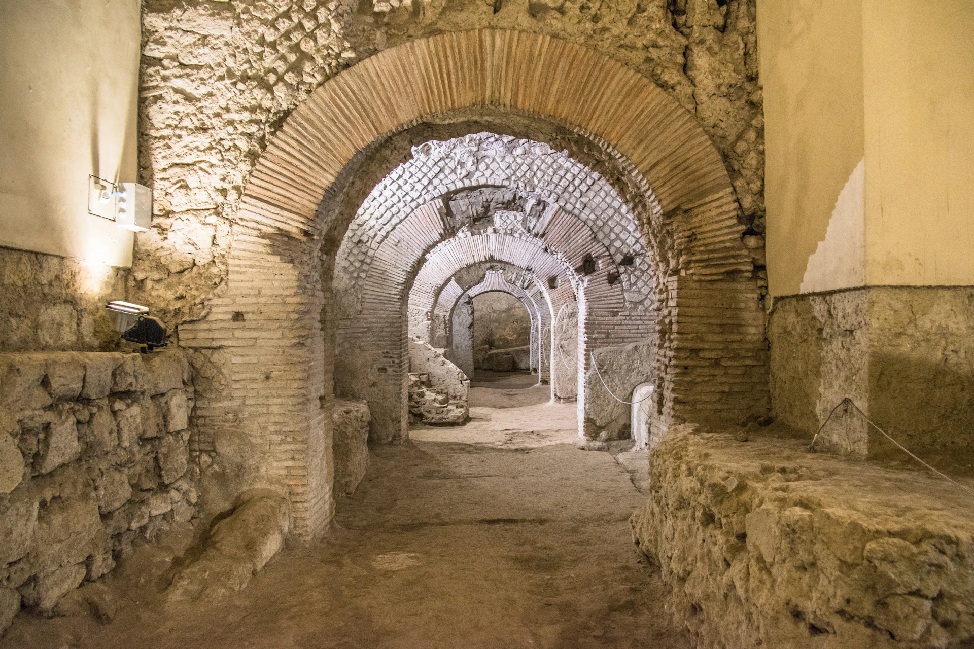 Ancient stone ruins with multiple arches, brick and stone masonry, dusty floor, and ambient light highlighting the structure.