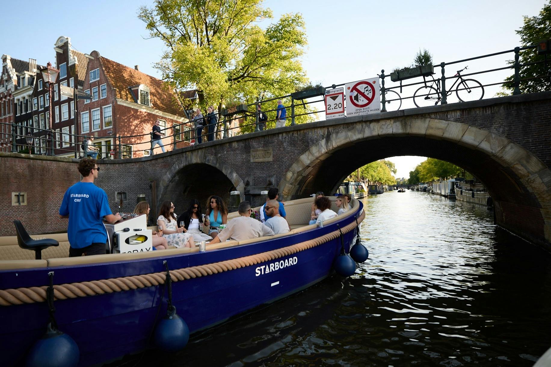 右舷という名前の青いボートに乗った人々のグループが、木や建物を背景にした運河の橋の下を通過しています。