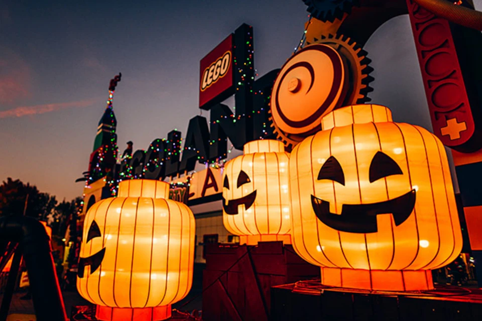 Glowing pumpkin lanterns with smiling faces in front of a LEGO sign and colorful lights at dusk.