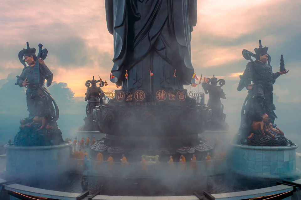 Large statue with smaller statues around it, surrounded by fog. Monks in orange robes positioned around the base. Cloudy sky at sunset.