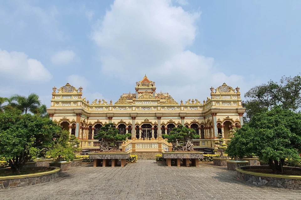Ornate yellow and gold temple with arched windows and intricate details, surrounded by manicured plants under a partly cloudy sky.