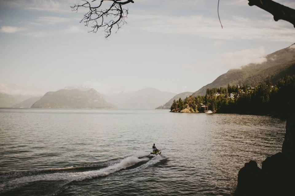 A person rides a jet ski on a calm lake surrounded by mountains and trees under a cloudy sky.