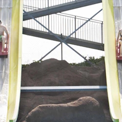 Två personer rider längs parallella vattenrutschbanor, var och en sitter på röda kuddar. Bakgrunden inkluderar bergformationer och en metalltrappa.